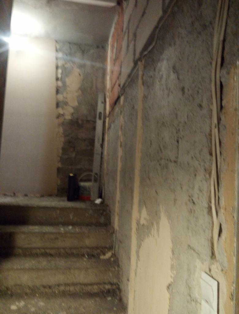 Klatka schodowa w trakcie remontu: surowe ściany z pozostałościami tynku, widoczne przewody elektryczne, poziomica oparta o ścianę, wiadro i pojemnik na schodach, przygotowanie do dalszych prac...