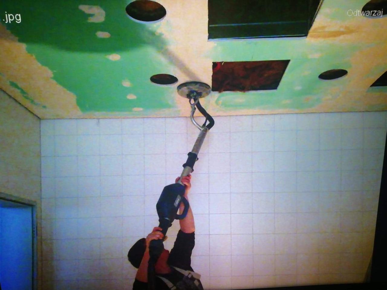 Szlifierka do gipsu na wysięgniku w trakcie pracy przy suficie z otworami na oświetlenie i częściowo pomalowanym na zielono, ściana wyłożona białymi płytkami.