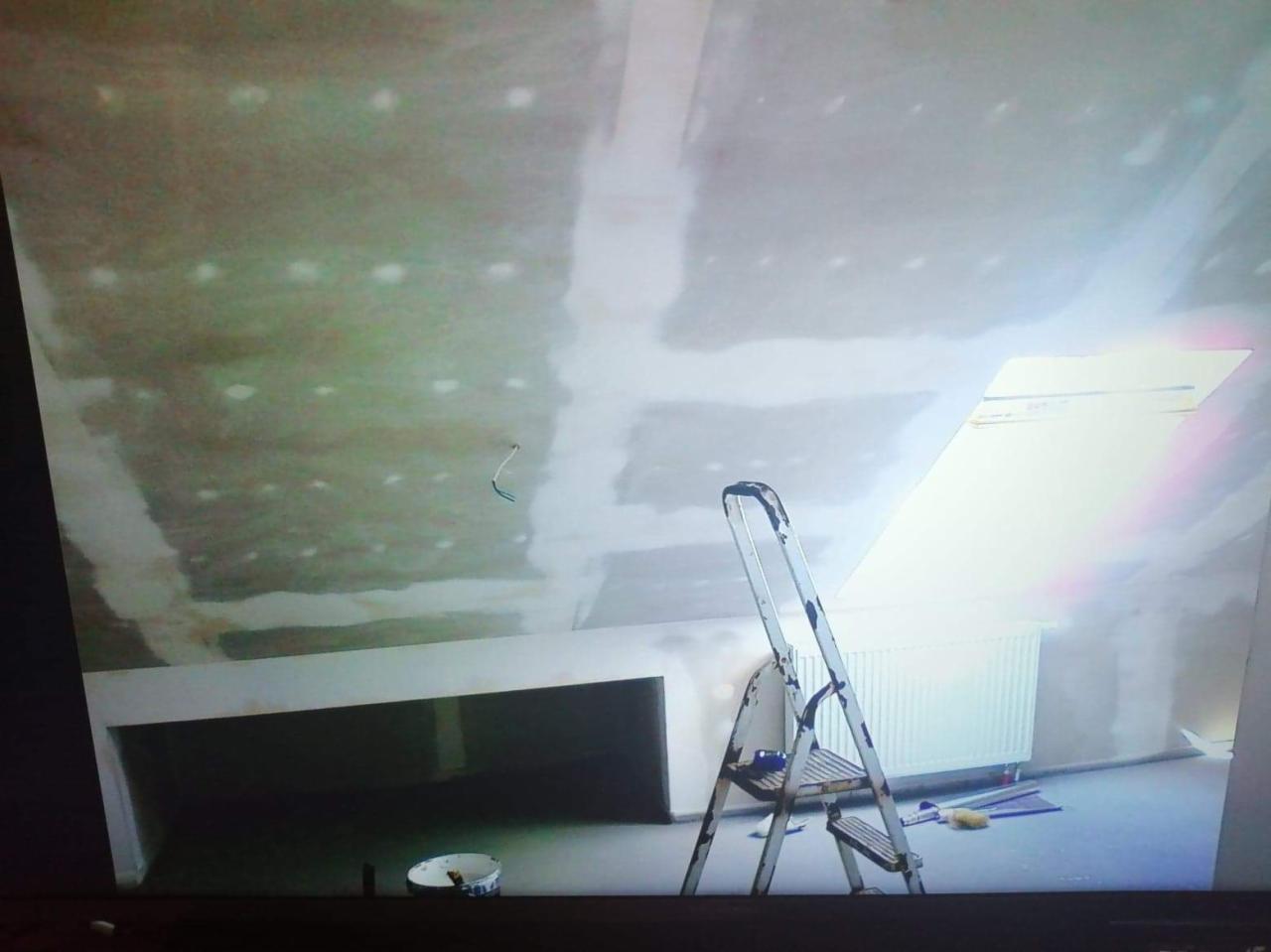 Poddasze w trakcie prac wykończeniowych, widoczne płyty kartonowo-gipsowe na suficie i ścianach, szpachlowane łączenia, drabina aluminiowa, wiadro z farbą i narzędzia malarskie na podłodze, okno...