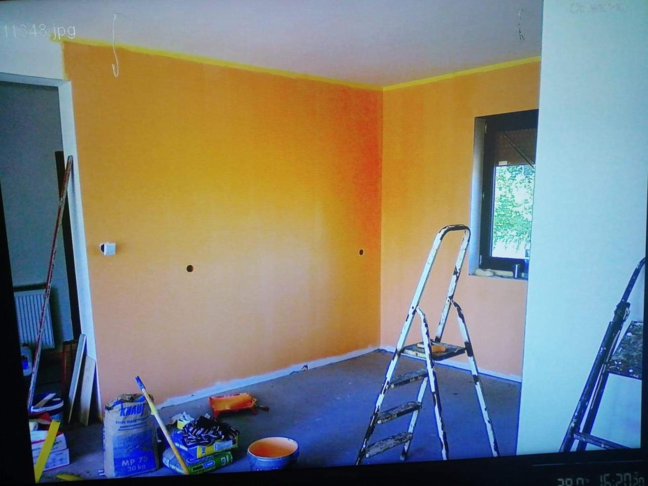 Pokój w trakcie malowania na pomarańczowo-żółty odcień, widoczna drabina, wiadro z farbą i worek z gładzią Knauf MP 75, zabezpieczona taśmą krawędź sufitu, przygotowane otwory pod gniazdka.