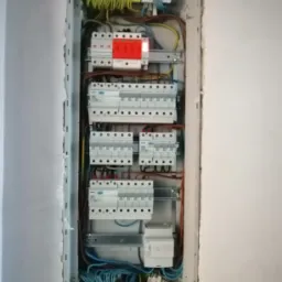 Otwarta skrzynka elektryczna w ścianie, z widocznymi bezpiecznikami, przewodami w kolorach brązowym, żółtym i niebieskim, oraz listwami zaciskowymi w kolorach zielonym i niebieskim.