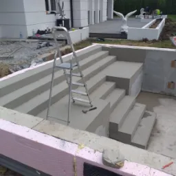 Betonowe schody z izolacją termiczną, drabina i młotek w tle domu z rzeźbami ptaków. Etap budowy z widocznymi materiałami budowlanymi.