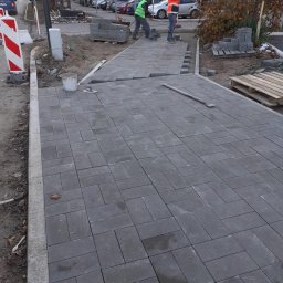 KOSTKABUD ŁUKASZ KOSTKA - Szara kostka brukowa ułożona na chodniku w jodełkę, z dwoma pracownikami w odblaskowych ubraniach układającymi kolejne elementy. Widoczny fragment ulicy i budynki w tle.