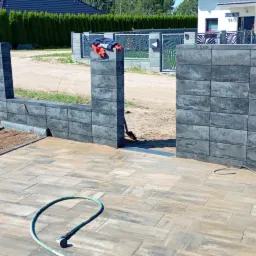 Nowoczesny taras z kostki brukowej w odcieniach beżu i szarości, z fragmentem muru z bloków betonowych w tle. Wąż ogrodowy leży na kostce.