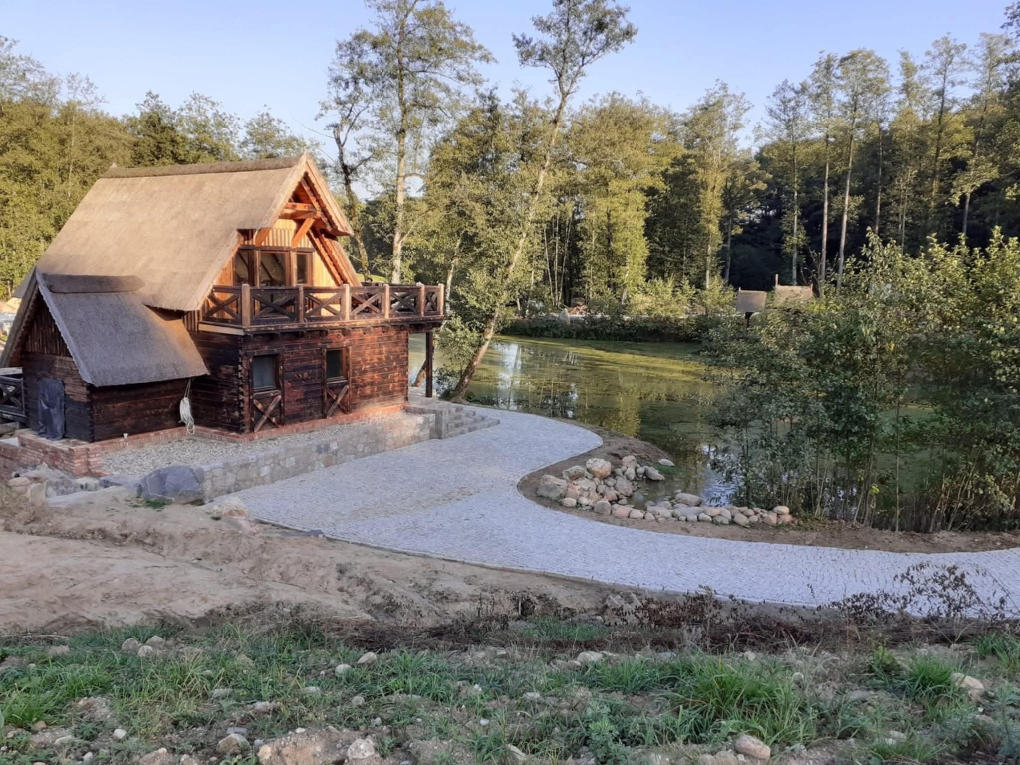 Drewniany dom z bali z gontowym dachem i ścieżką z jasnej kostki granitowej prowadzącą do stawu. Otoczenie leśne, widok z lekkiej góry.