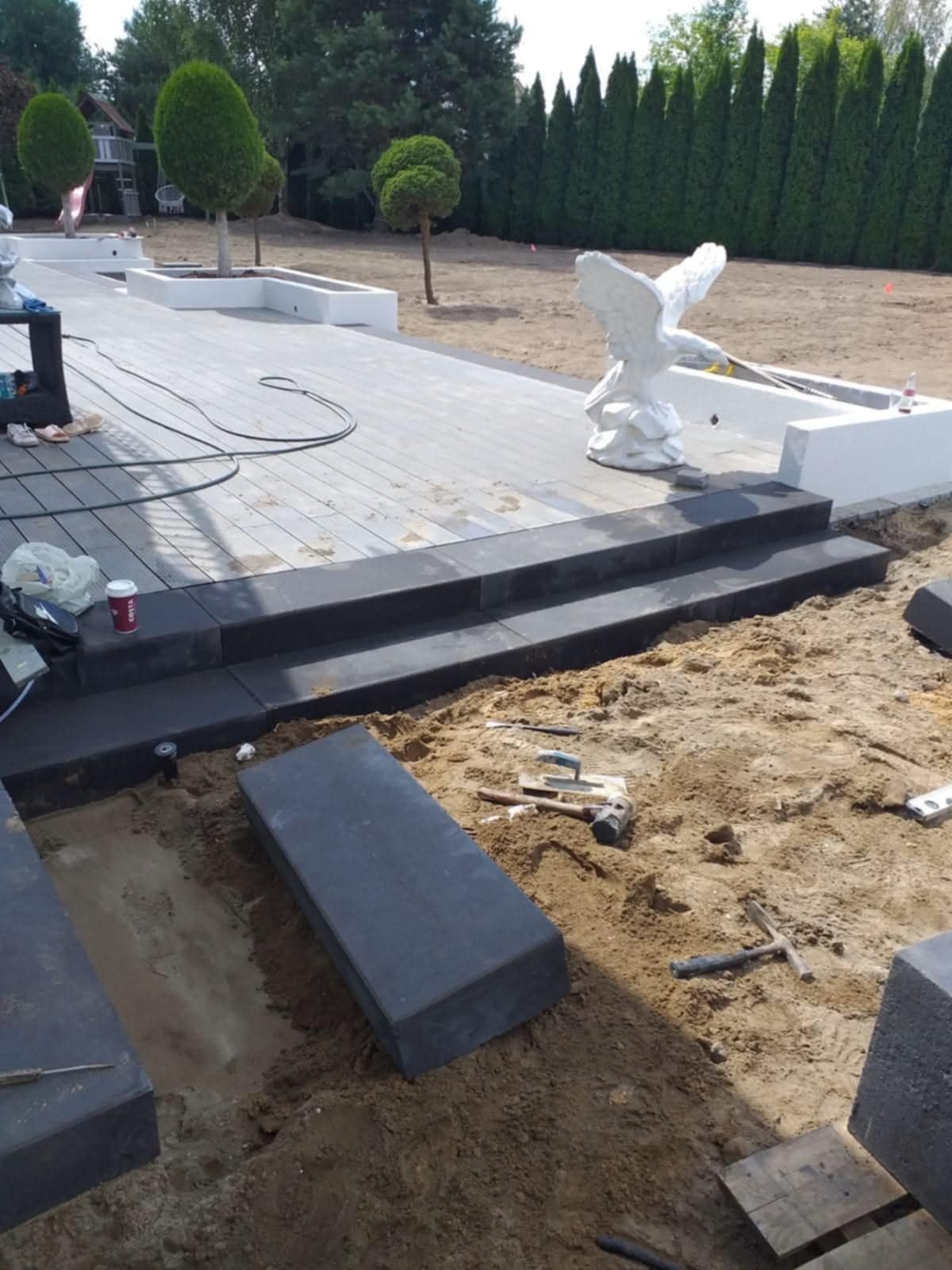 Ciemnoszare schody z kostki brukowej prowadzą na taras z jasnoszarej deski kompozytowej; w tle drzewa i rzeźba orła, widoczne narzędzia brukarskie.