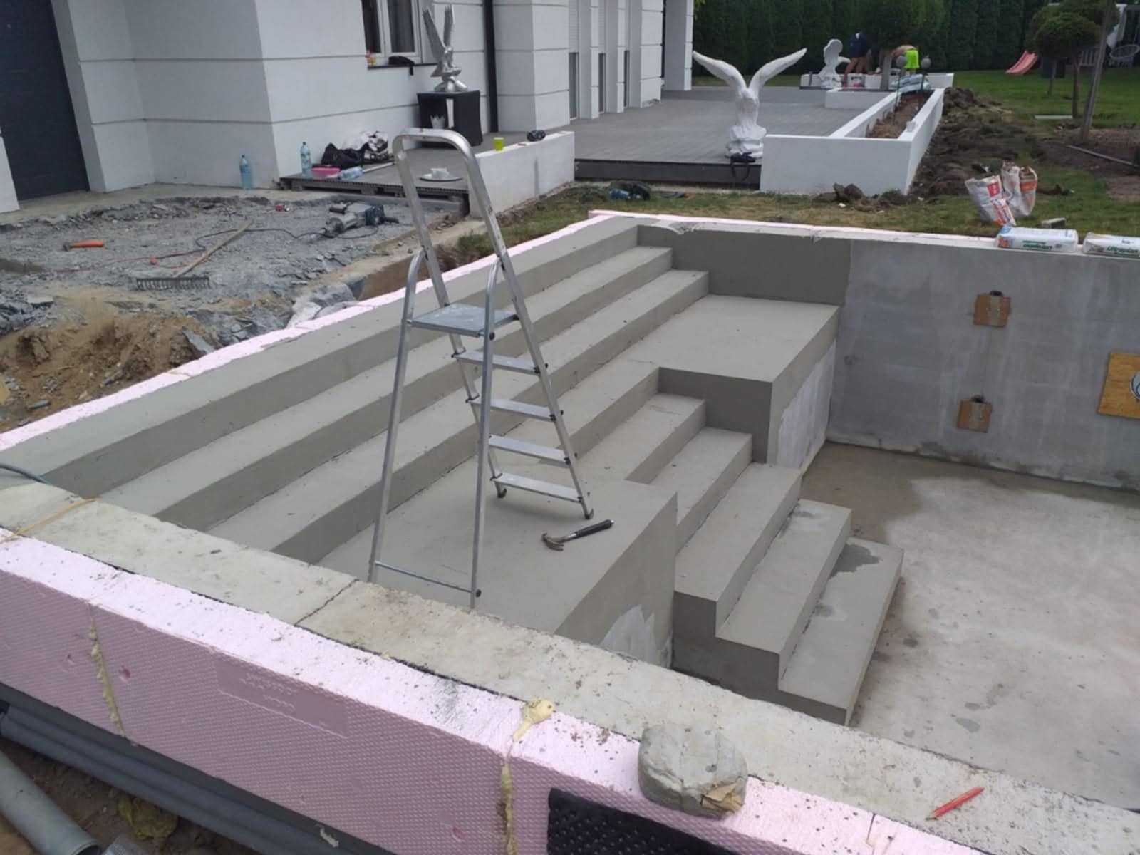 Betonowe schody z izolacją termiczną, drabina i młotek w tle domu z rzeźbami ptaków. Etap budowy z widocznymi materiałami budowlanymi.