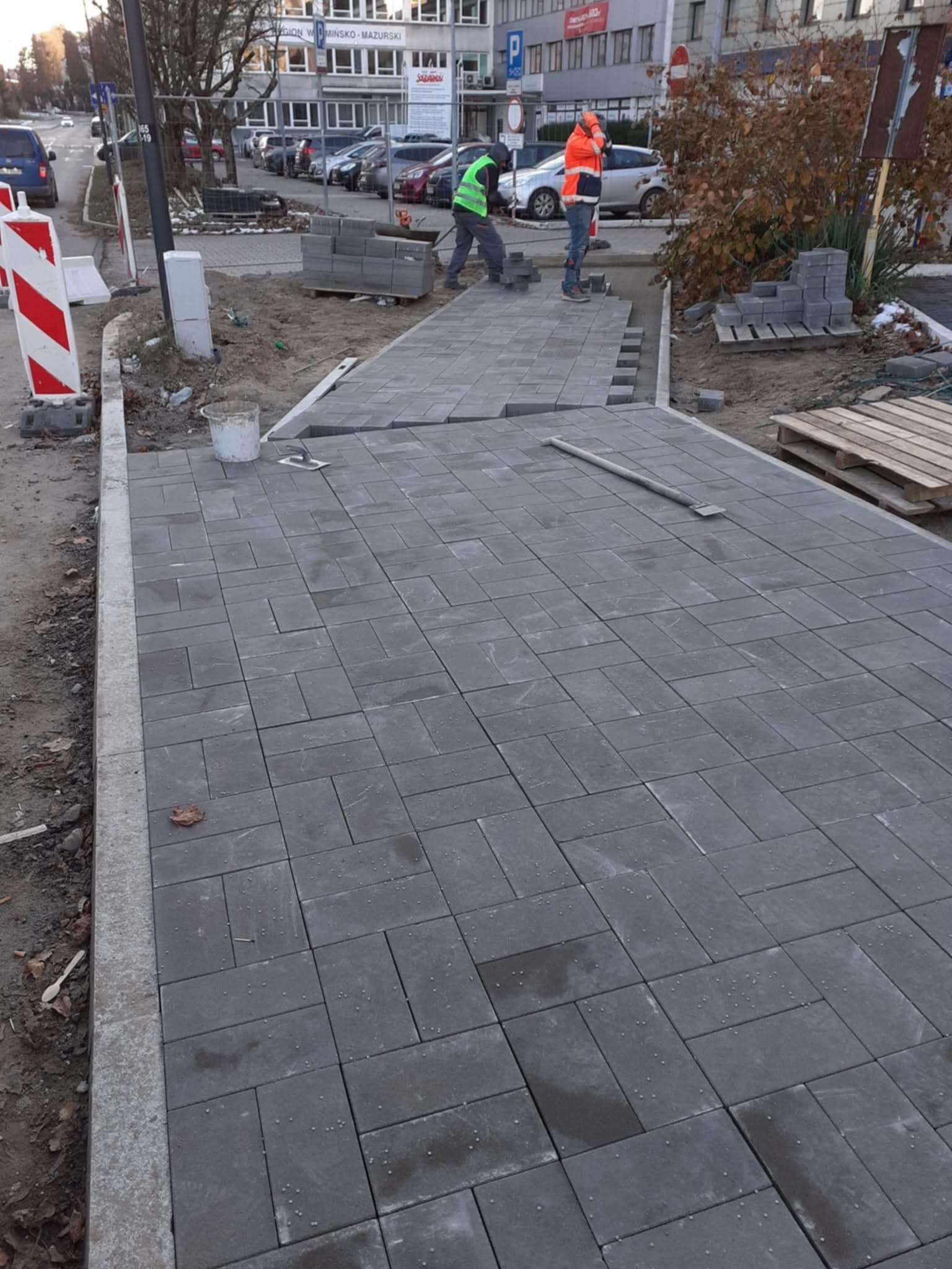 Szara kostka brukowa ułożona na chodniku w jodełkę, z dwoma pracownikami w odblaskowych ubraniach układającymi kolejne elementy. Widoczny fragment ulicy i budynki w tle.