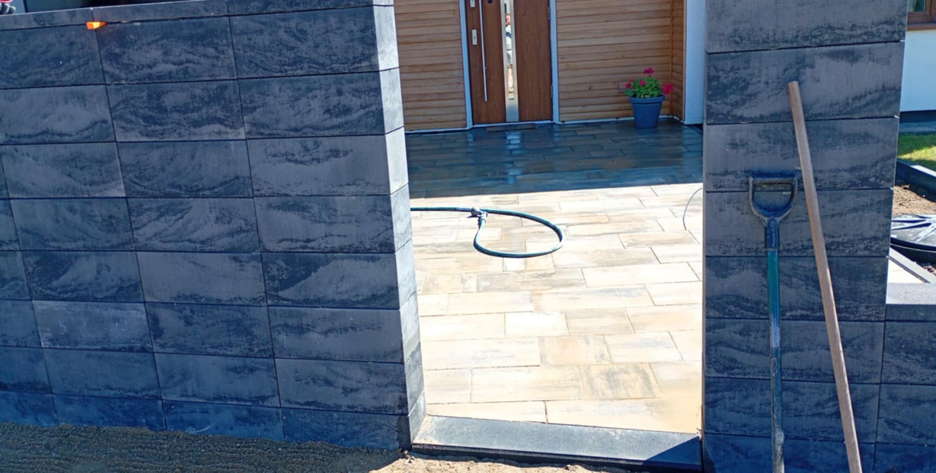 Nowo ułożona, beżowo-szara kostka brukowa, widoczna przez bramę z szarej kostki. W tle drewniany dom i doniczka z kwiatami. Obok ściany oparta łopata.