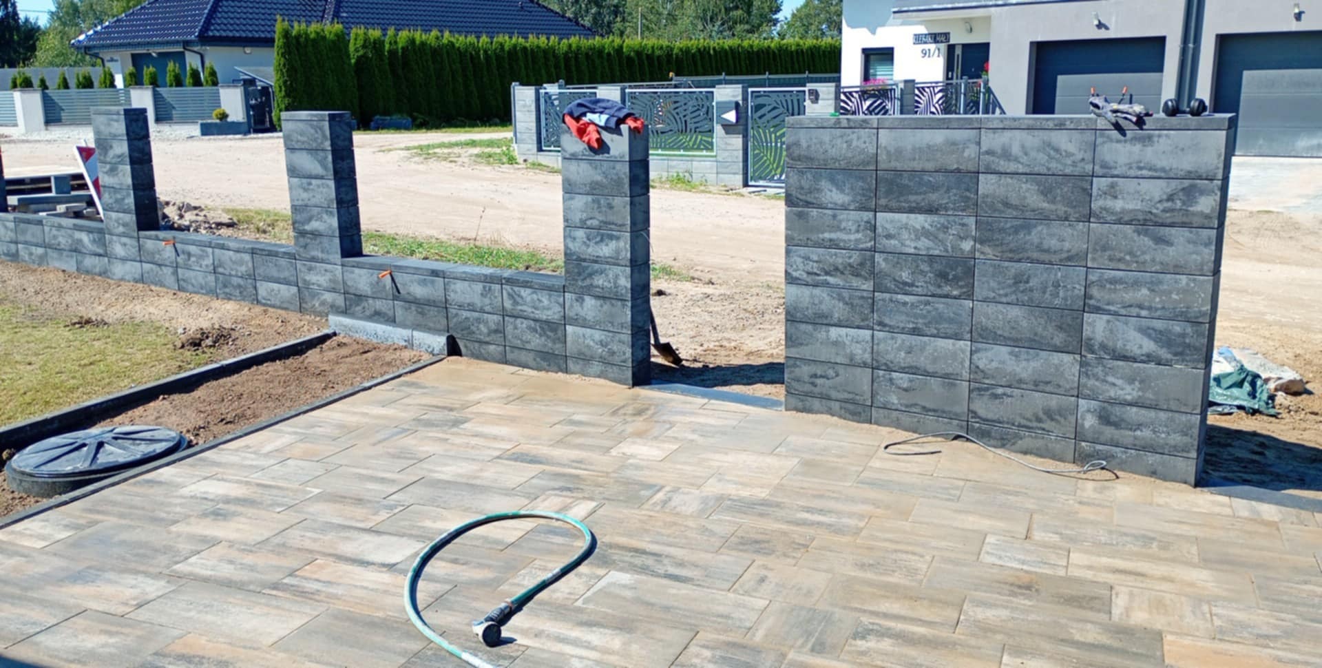 Nowoczesny taras z kostki brukowej w odcieniach beżu i szarości, z fragmentem muru z bloków betonowych w tle. Wąż ogrodowy leży na kostce.