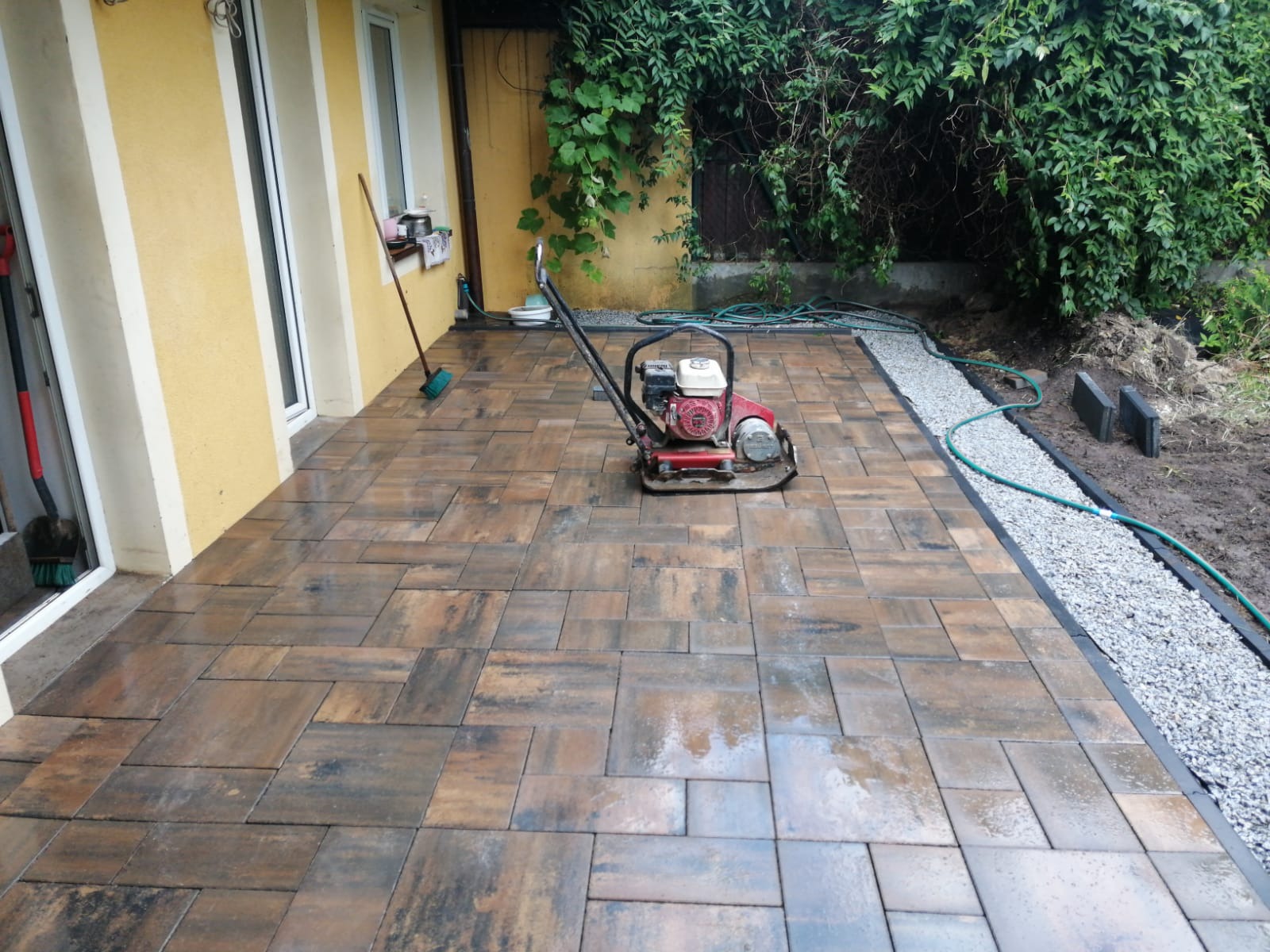 Ułożona kostka brukowa w odcieniach brązu, z zagęszczarką w centrum. Widoczny fragment ogrodu z zielenią i obrzeżem z drobnego kamienia. Żółta elewacja budynku w tle.