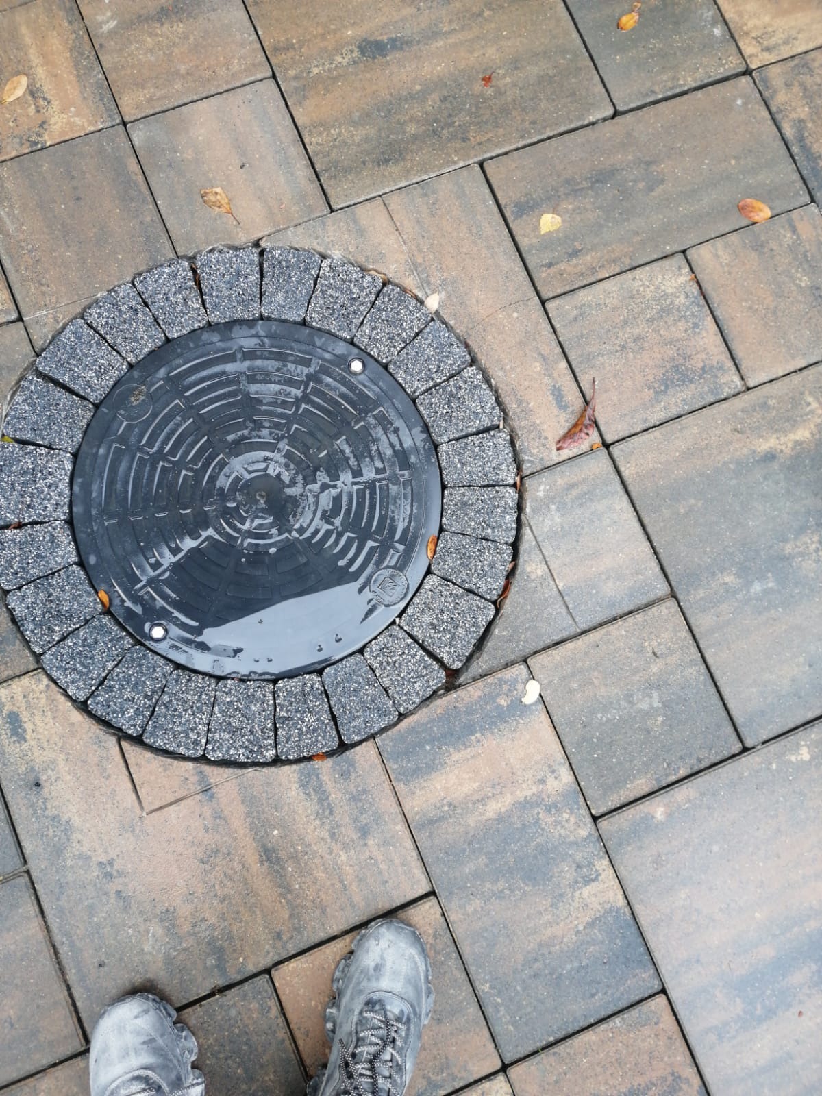 Estetyczne wykończenie studzienki kanalizacyjnej kostką brukową w odcieniach brązu i szarości, uzupełnionej o dekoracyjny okrąg z drobnej kostki.