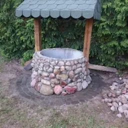 Kamienna studnia z drewnianą konstrukcją zadaszenia, otoczona kostką brukową i stertą kamieni, na tle zielonych drzew.