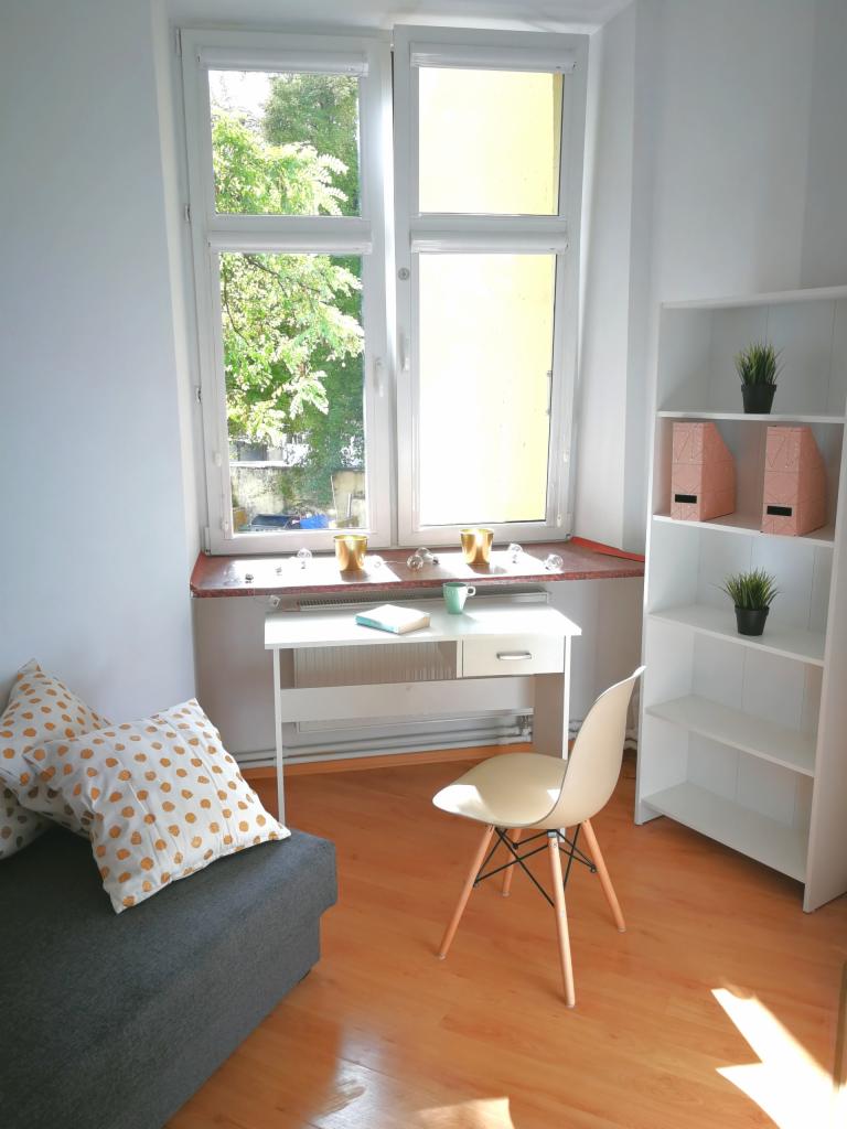 Jasny pokój z białym biurkiem, krzesłem Eames, szarą sofą z poduszkami w złote kropki i regałem z dekoracjami, widok z okna na zieleń.