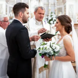 Para młoda podczas ceremonii ślubnej w kościele, ksiądz czyta z księgi, panna młoda trzyma bukiet z białych kwiatów.
