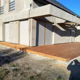 Surowy dom w trakcie wykończenia z nowym drewnianym tarasem, widoczny cień fotografa na pierwszym planie.