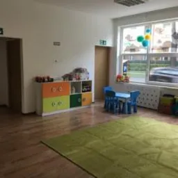 Jasne wnętrze sali zabaw z zielonym dywanem, kolorową komodą z zabawkami, stolikiem i krzesełkami dla dzieci, widokiem na ulicę przez okno i znakami ewakuacyjnymi.