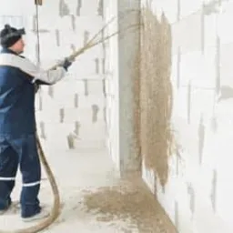 Pracownik w kombinezonie i ochronnych okularach natryskuje zaprawę na ścianę z bloczków betonowych za pomocą węża i dyszy pod ciśnieniem.