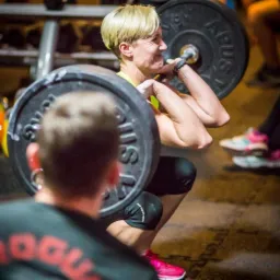 Kobieta z krótkimi blond włosami wykonuje przysiad ze sztangą w siłowni, widoczny grymas wysiłku, w tle inny mężczyzna i buty sportowe innej osoby.