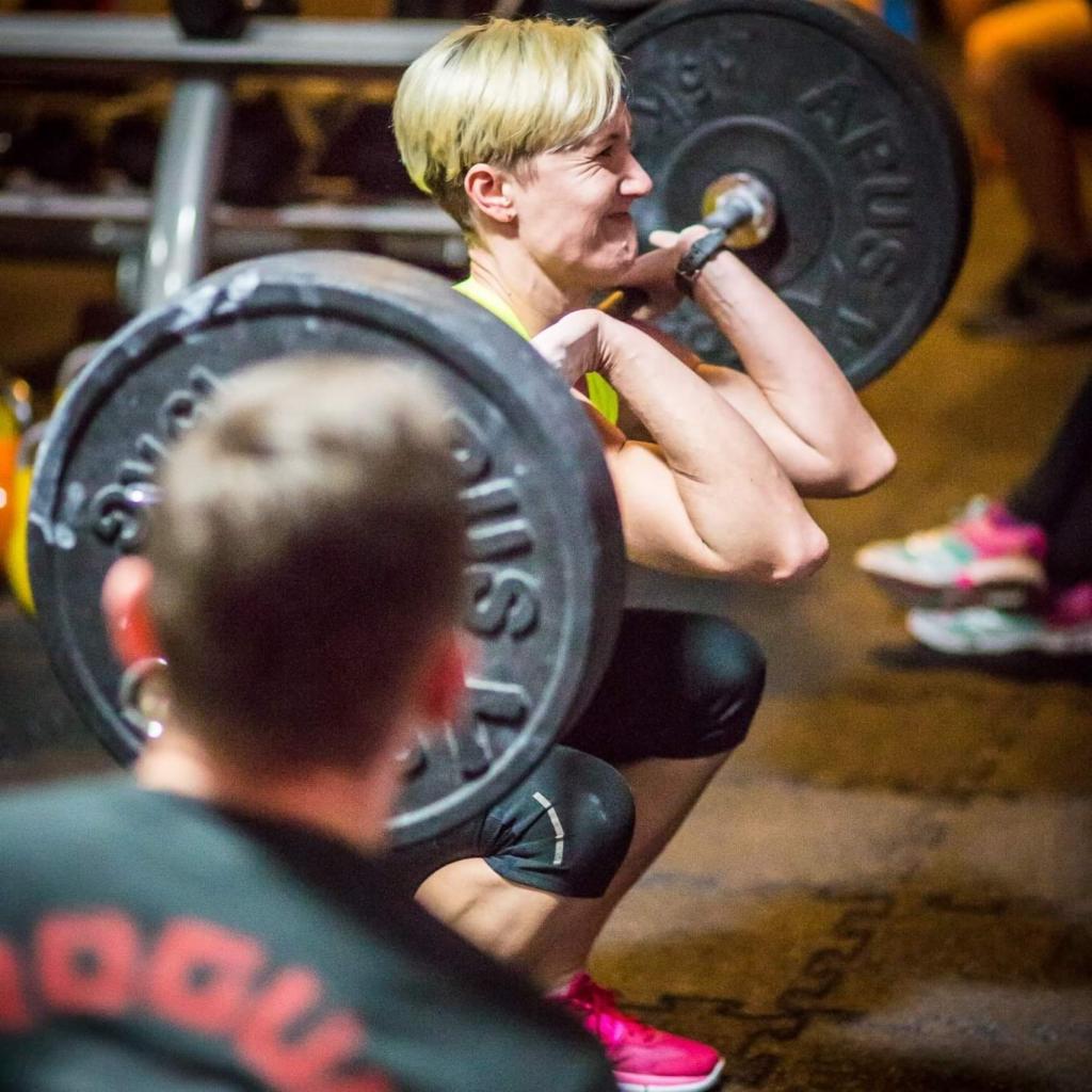 Kobieta z krótkimi blond włosami wykonuje przysiad ze sztangą w siłowni, widoczny grymas wysiłku, w tle inny mężczyzna i buty sportowe innej osoby.