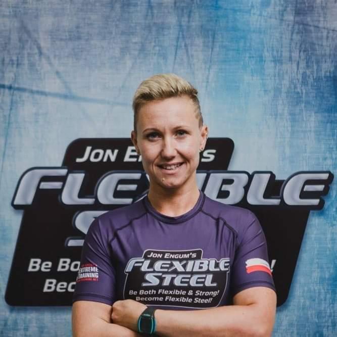 Kobieta z krótkimi blond włosami, uśmiechnięta, w sportowej koszulce z napisem 'Flexible Steel', skrzyżowane ramiona, na tle z logo.