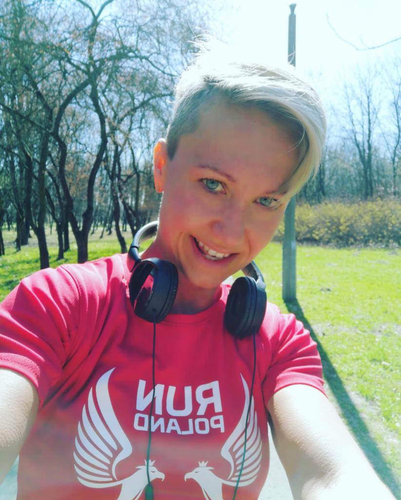 Uśmiechnięta blondynka z krótkimi włosami w czerwonej koszulce z nadrukiem 'RUN POLAND' i słuchawkami na szyi, robiąca selfie w parku.