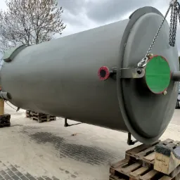 Duży, cylindryczny zbiornik przemysłowy w kolorze khaki, podwieszony na łańcuchach i umieszczony na paletach. Widoczne zabezpieczenia otworów w postaci czerwonych i zielonych osłon. W tle budynki...