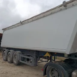 Biała naczepa wywrotka z plandeką, zaparkowana na placu, widok z boku na trzy osie i podwozie, pochmurne niebo w tle.