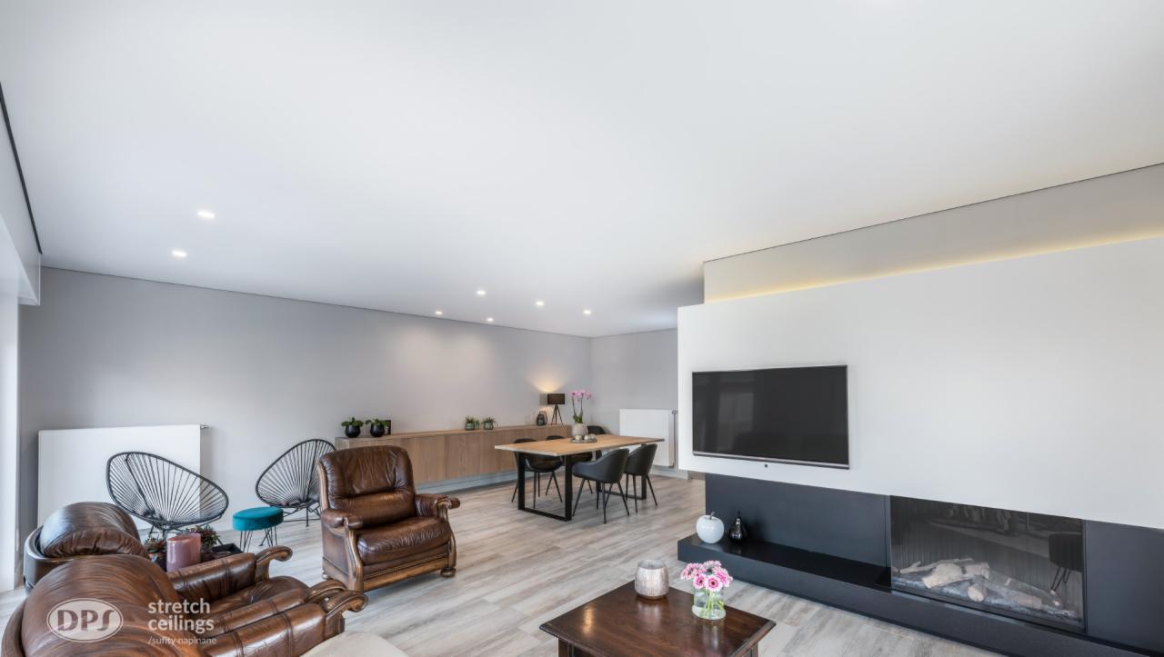 Jasne wnętrze salonu z białym sufitem napinanym z oświetleniem punktowym, brązowymi skórzanymi fotelami, drewnianym stołem i kominkiem z telewizorem