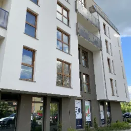 Nowoczesny budynek mieszkalny z balkonami i dużymi oknami, z widoczną reklamą lokali użytkowych na parterze i zadbaną zielenią przed wejściem.