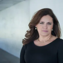 Portret kobiety w czarnej sukience, z perłowymi kolczykami i złotym naszyjnikiem. Ujęcie w jasnym, nowoczesnym wnętrzu z betonową ścianą w tle.