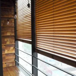 Drewniane żaluzje poziome w ciepłym odcieniu brązu, zamontowane w oknie z widokiem na miasto z balkonu wykończonego drewnem, oświetlone wiszącymi żarówkami bez klosza.