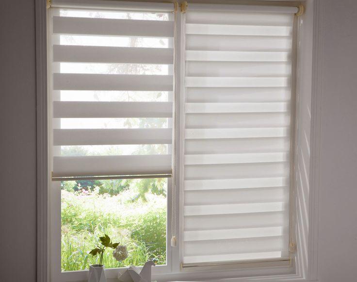 Okno z dwoma panelami, na jednym podniesiona, na drugim opuszczona, pozioma żaluzja z naprzemiennymi przezroczystymi i nieprzezroczystymi pasami, widok na zieleń za oknem, mała roślina w wazonie...