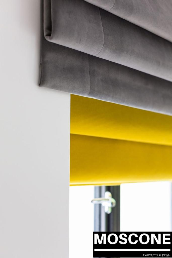 Szara, miękka tkanina ułożona w fałdy nad oknem z jaskrawożółtą przesłoną, widoczny fragment okna i logo firmy.