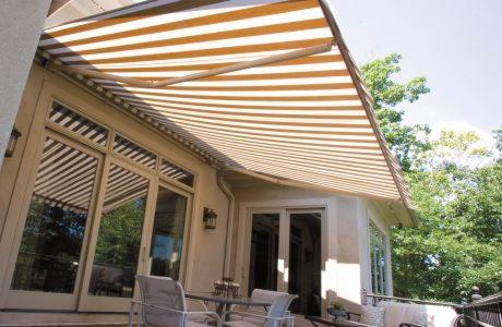 Beżowo-biała, rozkładana osłona przeciwsłoneczna nad tarasem z widokiem na zieleń.