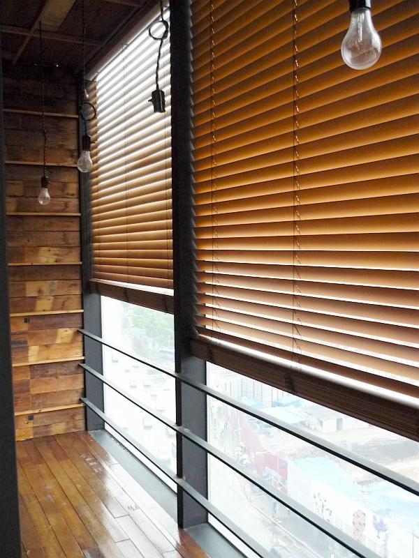 Drewniane żaluzje poziome w ciepłym odcieniu brązu, zamontowane w oknie z widokiem na miasto z balkonu wykończonego drewnem, oświetlone wiszącymi żarówkami bez klosza.