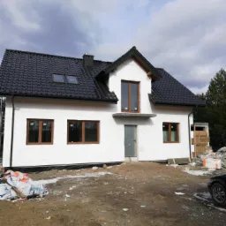 Surowy stan domu murowanego z białym styropianem elewacyjnym, ciemną dachówką i brązowymi oknami, widok z zewnątrz na plac budowy.