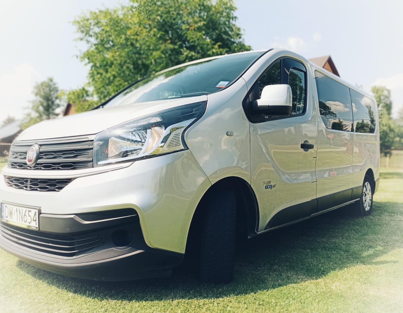 Srebrny bus Fiat z numerem rejestracyjnym DW 1N654, zaparkowany na zielonej trawie przed drzewami, widok z przodu z lekkiego dołu.