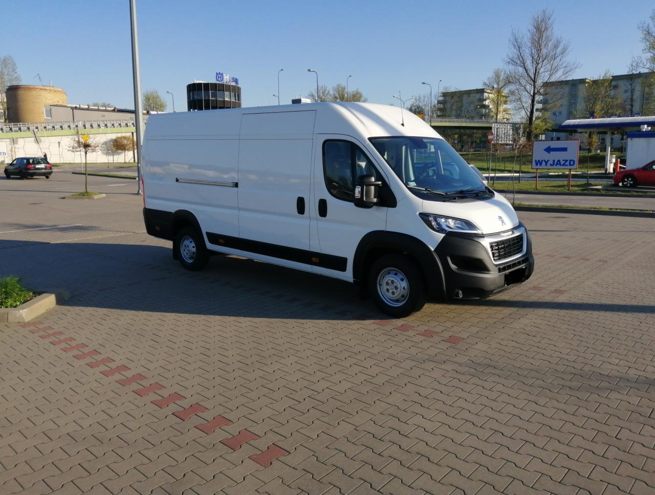 Biały bus Peugeot Boxer na parkingu z kostki brukowej, znak 'Wyjazd' w tle, widok z przodu i boku.
