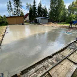 Wylany fundament z mokrego betonu, wyrównywany przez dwóch pracowników za pomocą specjalistycznej listwy w otoczeniu drewnianych szalunków i narzędzi.