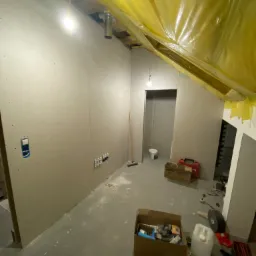 Wnętrze poddasza w trakcie wykańczania: ściany obłożone płytami gipsowo-kartonowymi z widocznymi śrubami, instalacja elektryczna w puszkach, zabezpieczona folią budowlaną podbitka, narzędzia...