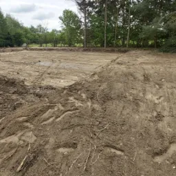 Przygotowany teren pod budowę z widocznymi śladami opon maszyn budowlanych i wyznaczonym obrysem przyszłego budynku, otoczony drzewami.