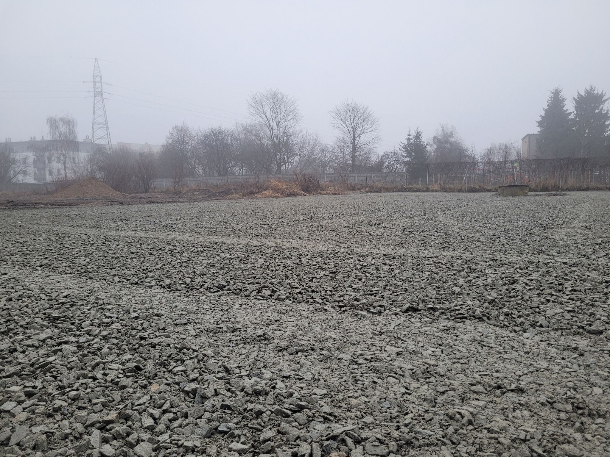 Szary tłuczeń na wyrównanym terenie z widoczną linią wysokiego napięcia i drzewami w tle, spowity mgłą.