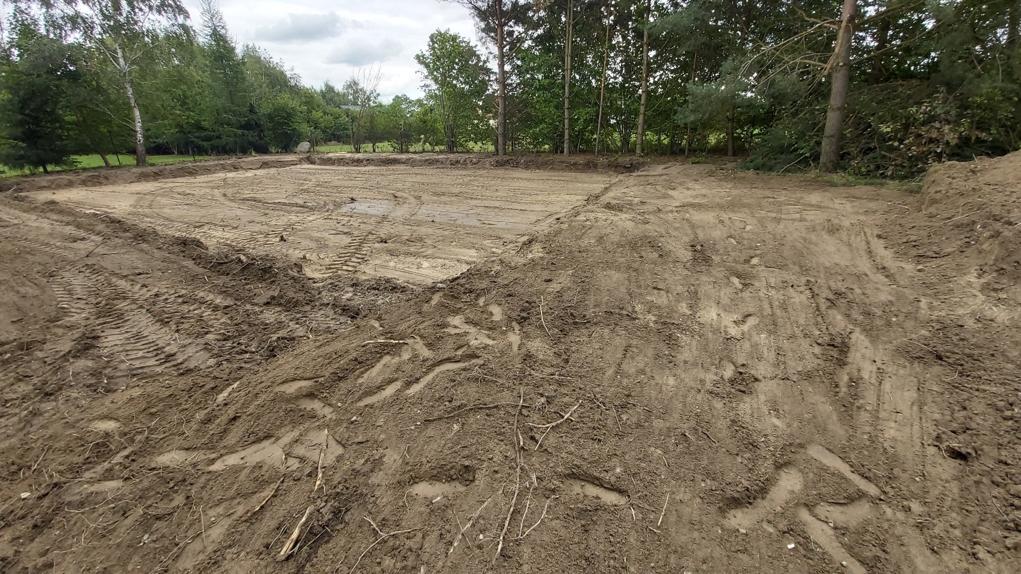 Przygotowany teren pod budowę z widocznymi śladami opon maszyn budowlanych i wyznaczonym obrysem przyszłego budynku, otoczony drzewami.
