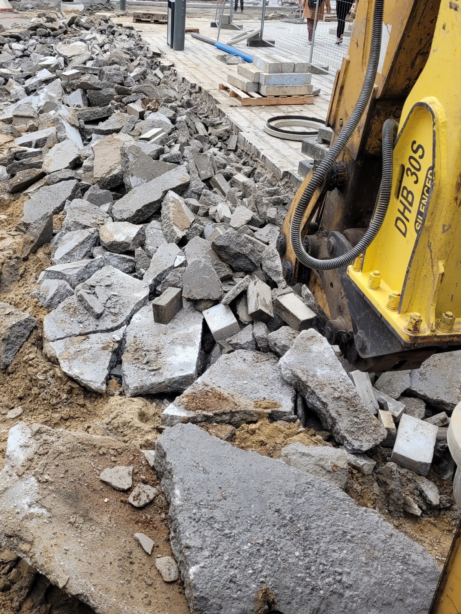 Żółty młot hydrauliczny DHB 30S kruszy betonową nawierzchnię na placu budowy; widoczne fragmenty chodnika z jasnej kostki i gruz.