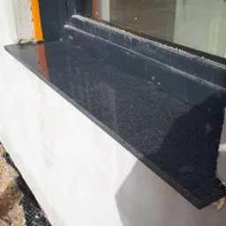 Czarny, błyszczący parapet z drobnego kamienia kompozytowego, zamontowany pod oknem w trakcie prac wykończeniowych elewacji.