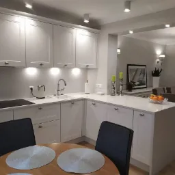 Jasna kuchnia z białymi szafkami i blatem, okrągły drewniany stół z szarymi krzesłami na pierwszym planie, widok na jadalnię w tle.