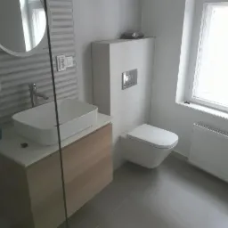 Nowoczesna łazienka z umywalką nablatową na szafce z drewnopodobnym frontem, podwieszaną toaletą i grzejnikiem pod oknem.