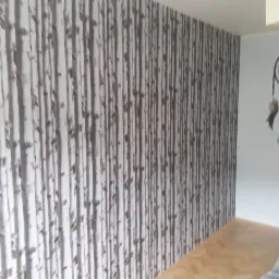 Ściana pokryta tapetą imitującą brzozowy las, z widocznym łapaczem snów wiszącym na ścianie w rogu pokoju.