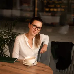 Kobieta w okularach siedzi przy okrągłym drewnianym stole z filiżanką herbaty i zielonym notatnikiem w kawiarni z roślinami doniczkowymi i odbiciem witryny w tle.